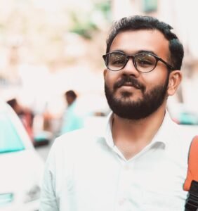 Confident man with glasses and backpack on a sunny urban street with cars in the background.